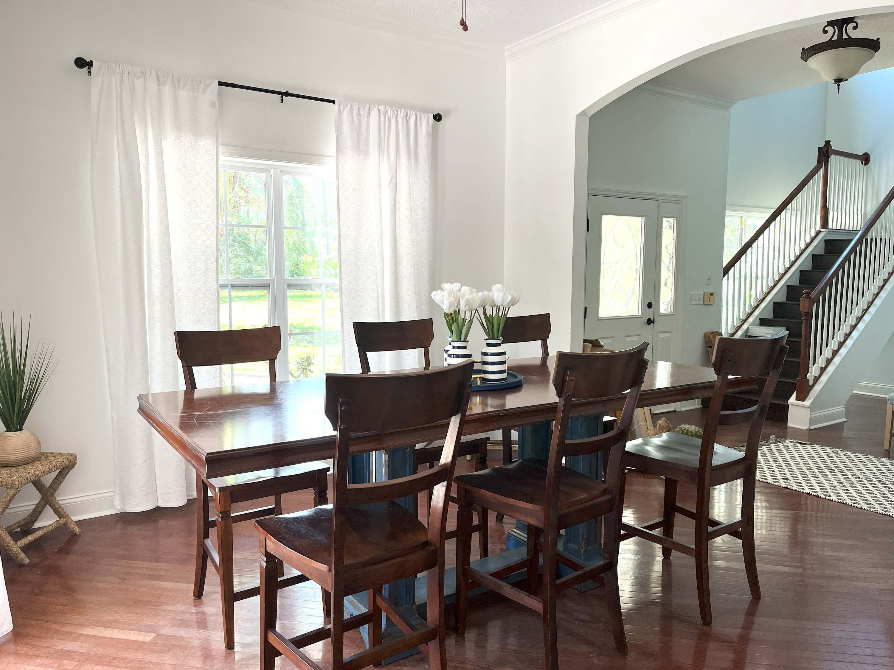 Un comedor con una mesa y sillas de madera, una planta en maceta y un ventanal con cortinas.
