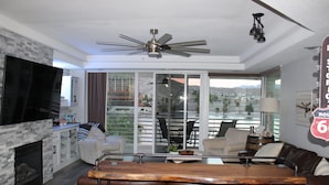 Un salon moderne doté d’une cheminée en pierre, d’un coin bar avec des tabourets, d’un ventilateur de plafond et offrant une vue sur un paysage côtier grâce à de grandes portes vitrées.