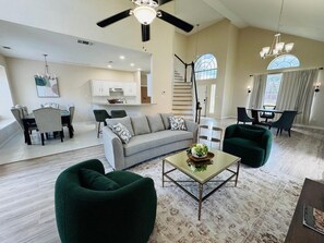 A spacious living room with a grey sofa, green armchairs, and a coffee table. A staircase leads to a kitchen area with white cabinets and a dining table. - Andalusia Trail Getaway (Arlington)