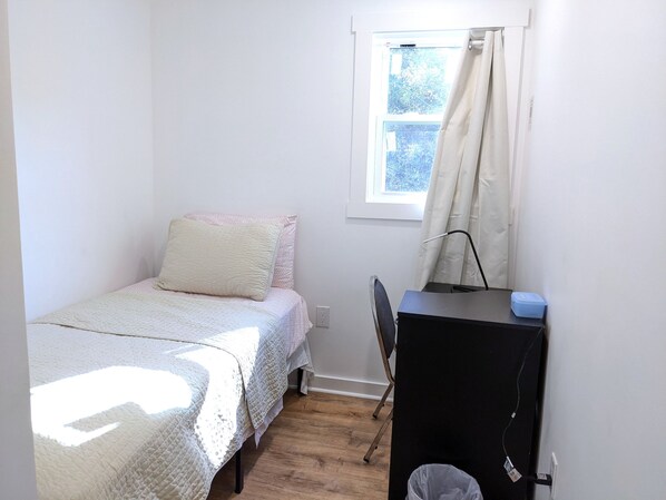 A small, single-bed room with a desk and chair, a window with curtains, and a waste bin.