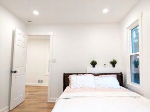 A bedroom with a bed, a window, a door, and a small plant on a shelf.