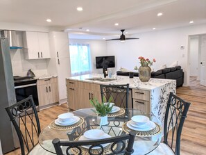 A modern kitchen with white cabinets, a dining area with a glass table, and a ceiling fan.