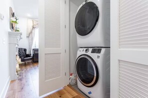 A laundry room with two stacked washing machines, a door leading to a living area with a chair and a small table, and a plant on the wall.
