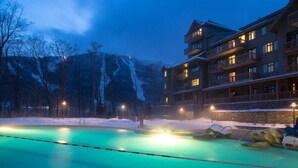 Un hotel con piscina, terreno cubierto de nieve y montañas al fondo.