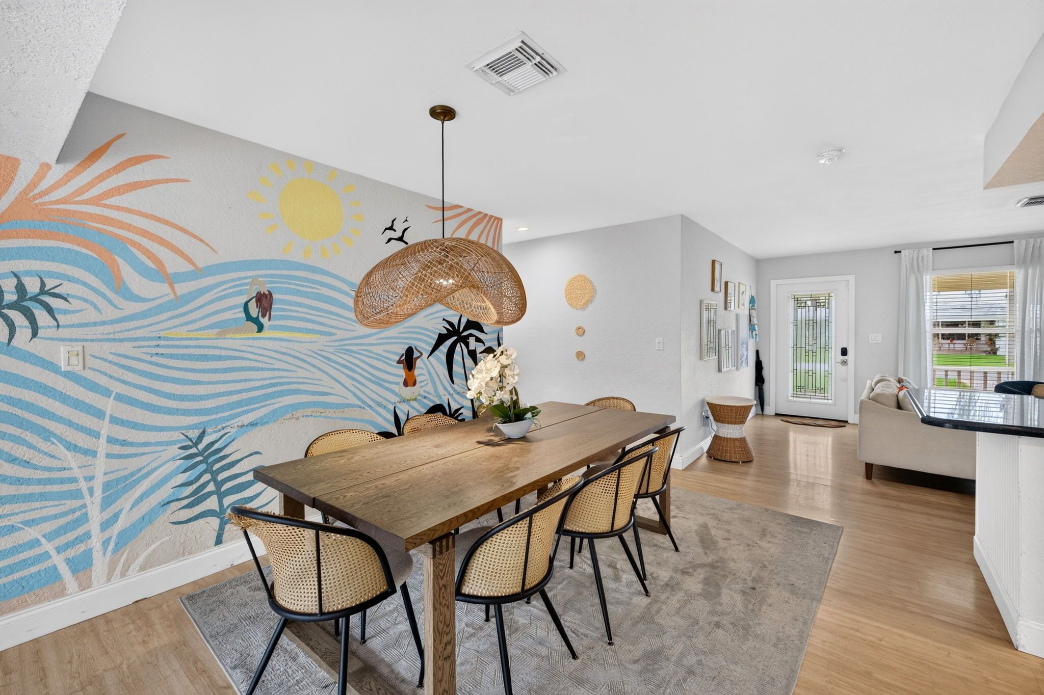 Un espace repas avec une table en bois, des chaises en osier et une fresque murale aux motifs tropicaux.