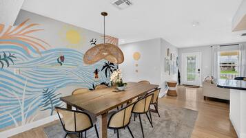 Un espace repas avec une table en bois, des chaises en osier et une fresque murale aux motifs tropicaux.