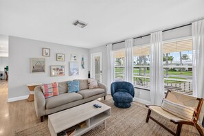 A living room with a sofa, coffee table, and a blue chair. There are framed pictures on the wall and a window with blinds.