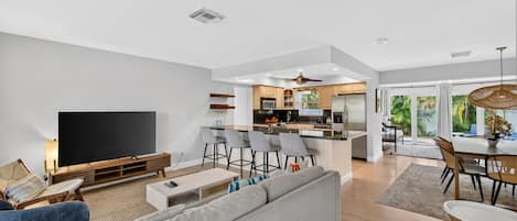 A modern living room with a flat-screen TV, a grey sofa, and a dining area with a table and chairs.