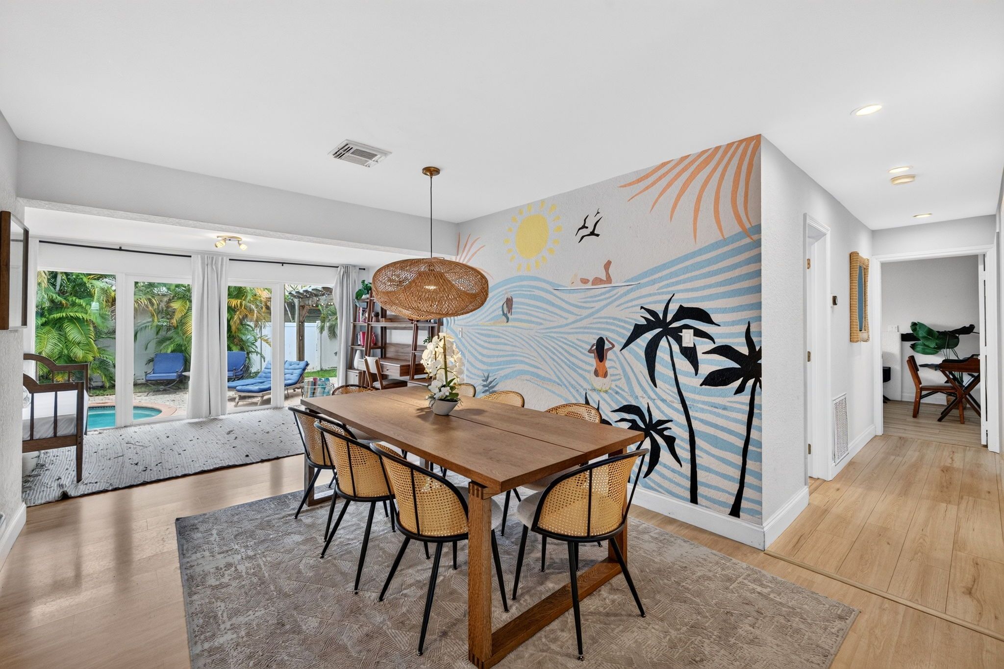Un espace repas avec une table et des chaises en bois, un luminaire suspendu en fibres tressées et une fresque murale aux motifs tropicaux.