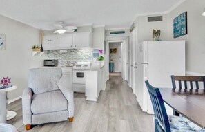 Cozinha compacta com armários brancos, micro-ondas e geladeira. Possui um espaço de refeições com mesa e cadeiras de madeira.