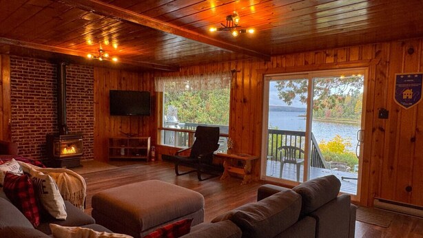 L’intérieur chaleureux d’un chalet avec une cheminée, des sièges confortables et une grande fenêtre donnant sur un lac.
