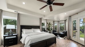 A bedroom with a large bed, a ceiling fan, a nightstand with a lamp, and a view of the outdoors through large windows. - Hoots Hollar Haus For 6 (Wimberley)