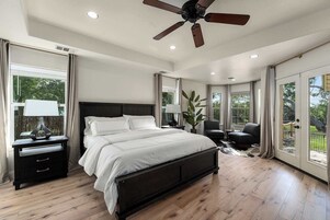 Un dormitorio con una cama grande, un ventilador de techo, una mesita de noche con lámpara y vistas al exterior a través de amplias ventanas.