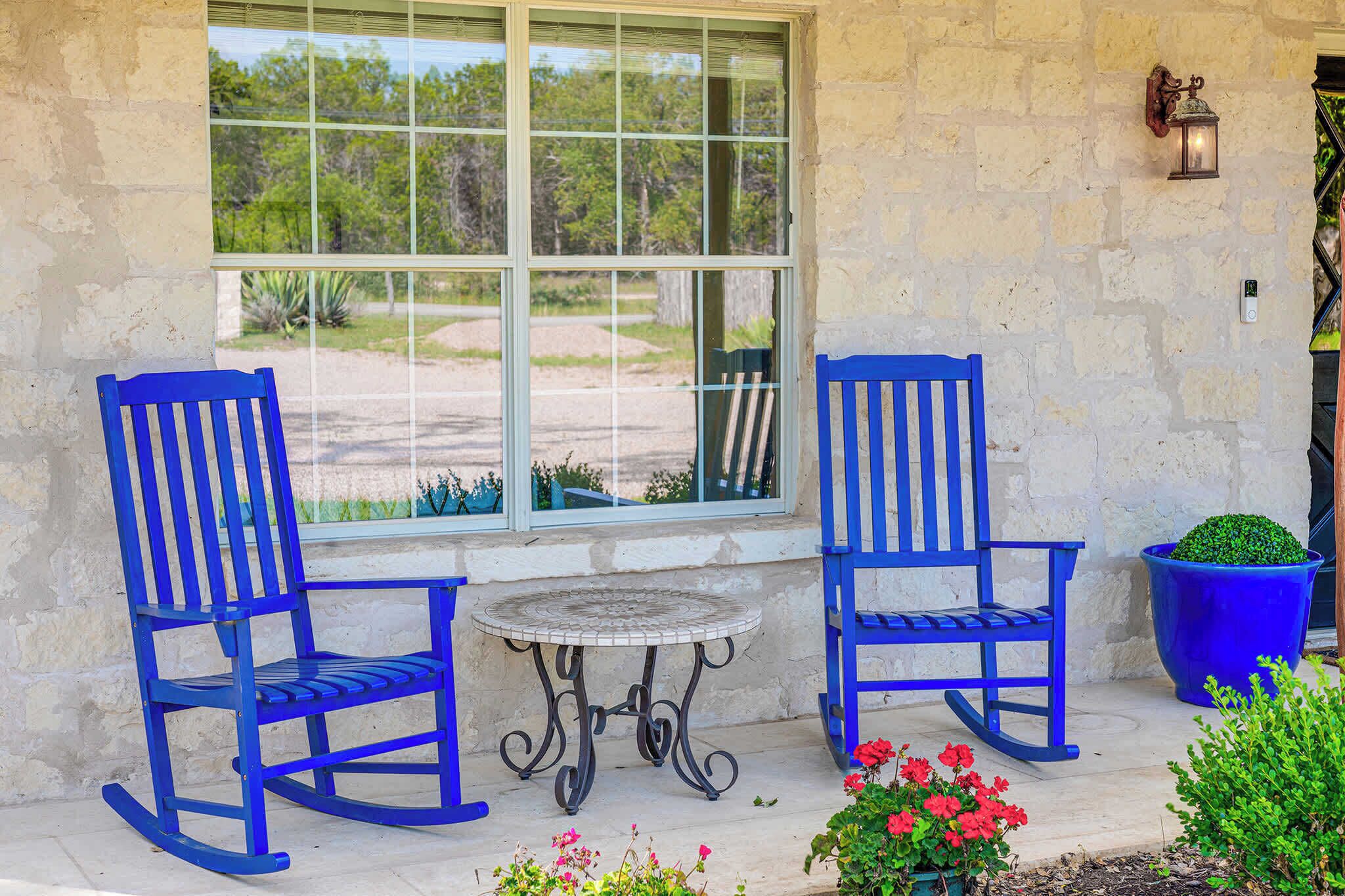 Two rocking chairs, a small table, and a potted plant on a patio with a stone wall and a window overlooking greenery.
