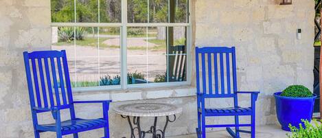 Two rocking chairs, a small table, and a potted plant on a patio with a stone wall and a window overlooking greenery.