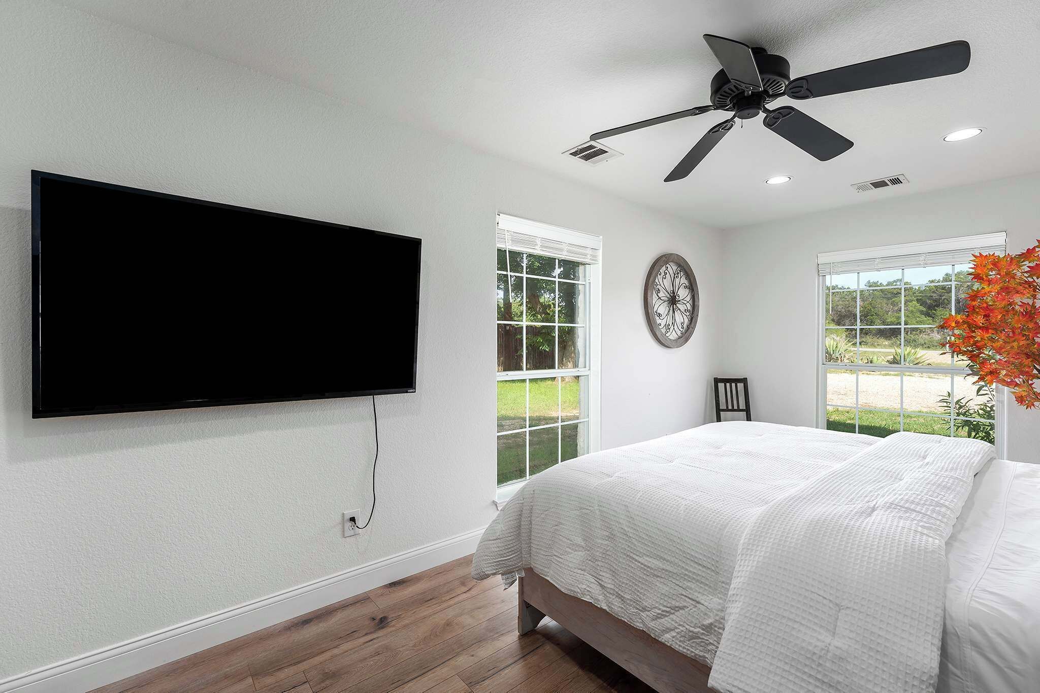 A bedroom with a bed, a ceiling fan, a TV mounted on the wall, and a window with a view of greenery.