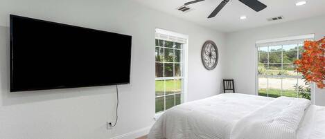 A bedroom with a bed, a ceiling fan, a TV mounted on the wall, and a window with a view of greenery.