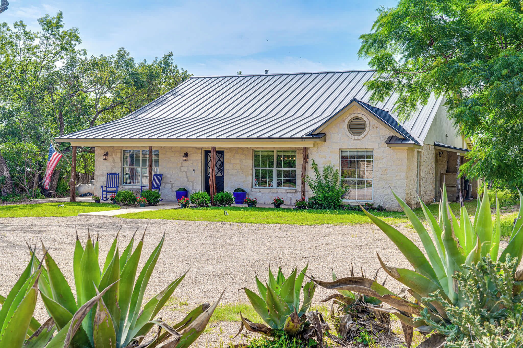 A single-story house with a stone facade, a metal roof, and a front porch with blue chairs.