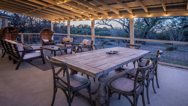 An outdoor patio with a wooden table and chairs, surrounded by wicker furniture and string lights. - Hoots Hollar Haus For 6 (Wimberley)