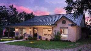 A single-story house with a metal roof, a front porch with chairs, and a flag. - Hoots Hollar Haus For 6 (Wimberley)