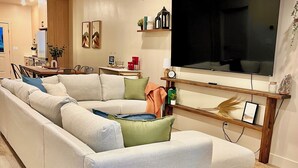 A modern living room with a large sectional sofa, a flat-screen TV, wooden shelving, and a dining area.