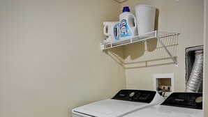 A laundry room with two front-loading washing machines, a shelf with cleaning supplies, and a vent on the wall.