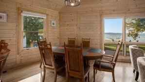 A wooden cabin interior with a round dining table and chairs, large windows with views of the outdoors, and a hanging light fixture.