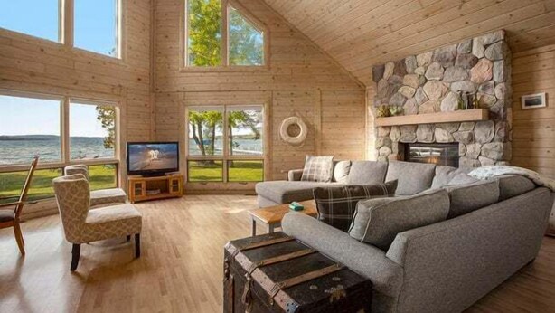 A cozy cabin interior with a stone fireplace, large windows overlooking a lake, and a comfortable seating area.