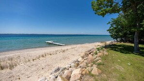 A beach with a wooden pier extending into the water, a grassy area with rocks, and a clear blue sky. - Old Mission Peninsula Luxury on West Bay (Traverse City)