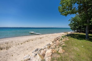A beach with a wooden pier extending into the water, a grassy area with rocks, and a clear blue sky.
