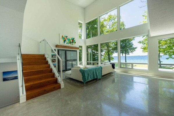 A spacious living area with a staircase, a sofa, and large windows overlooking a body of water.