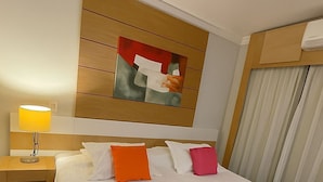 A hotel room with a large bed, a bedside table with a lamp, a painting on the wall, and a window with curtains.