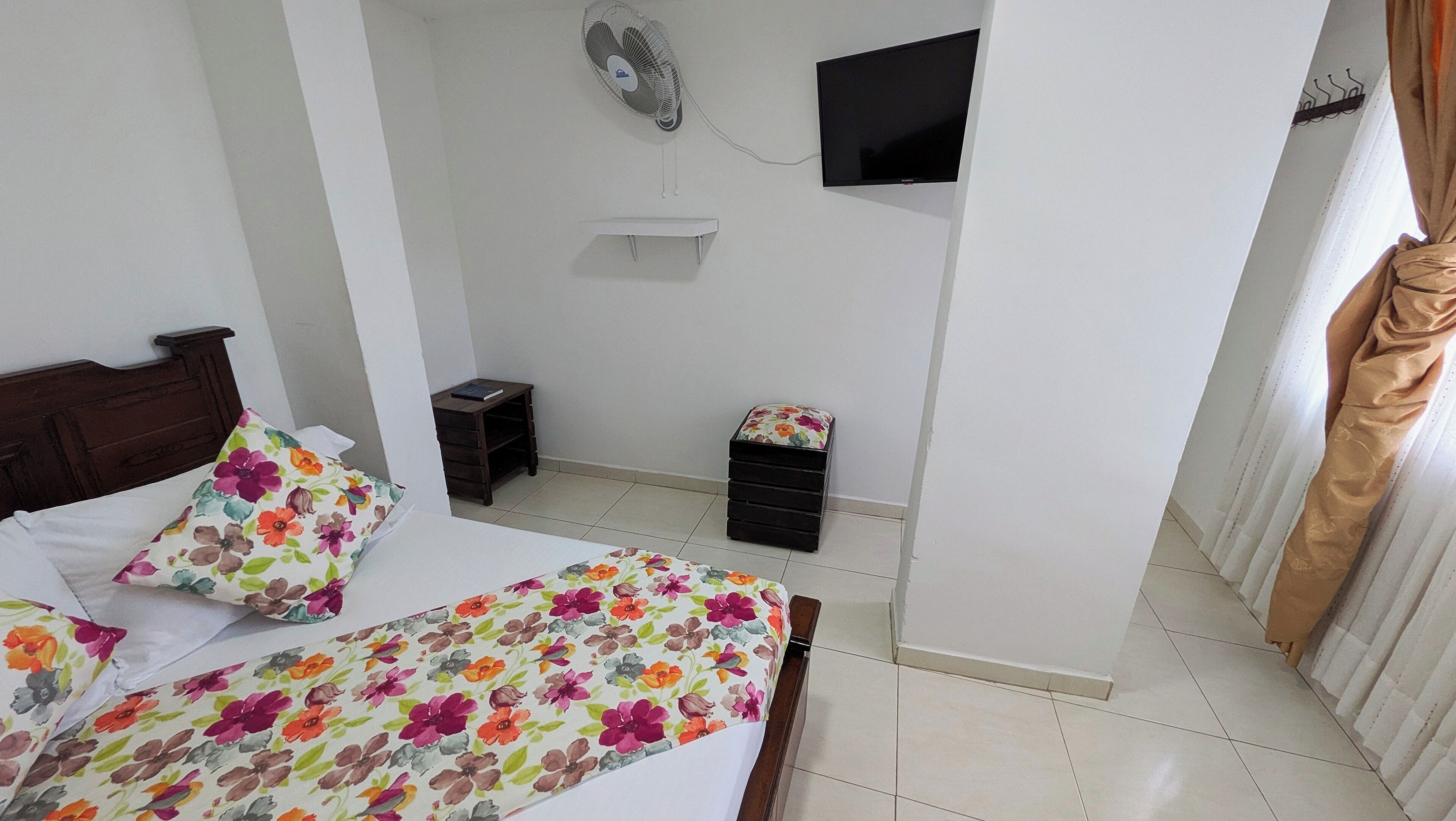 A bedroom with a bed, a small bedside table, a wall-mounted TV, and a standing fan.