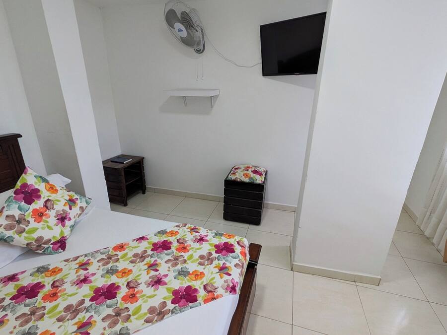 A bedroom with a bed, a small bedside table, a wall-mounted TV, and a standing fan.