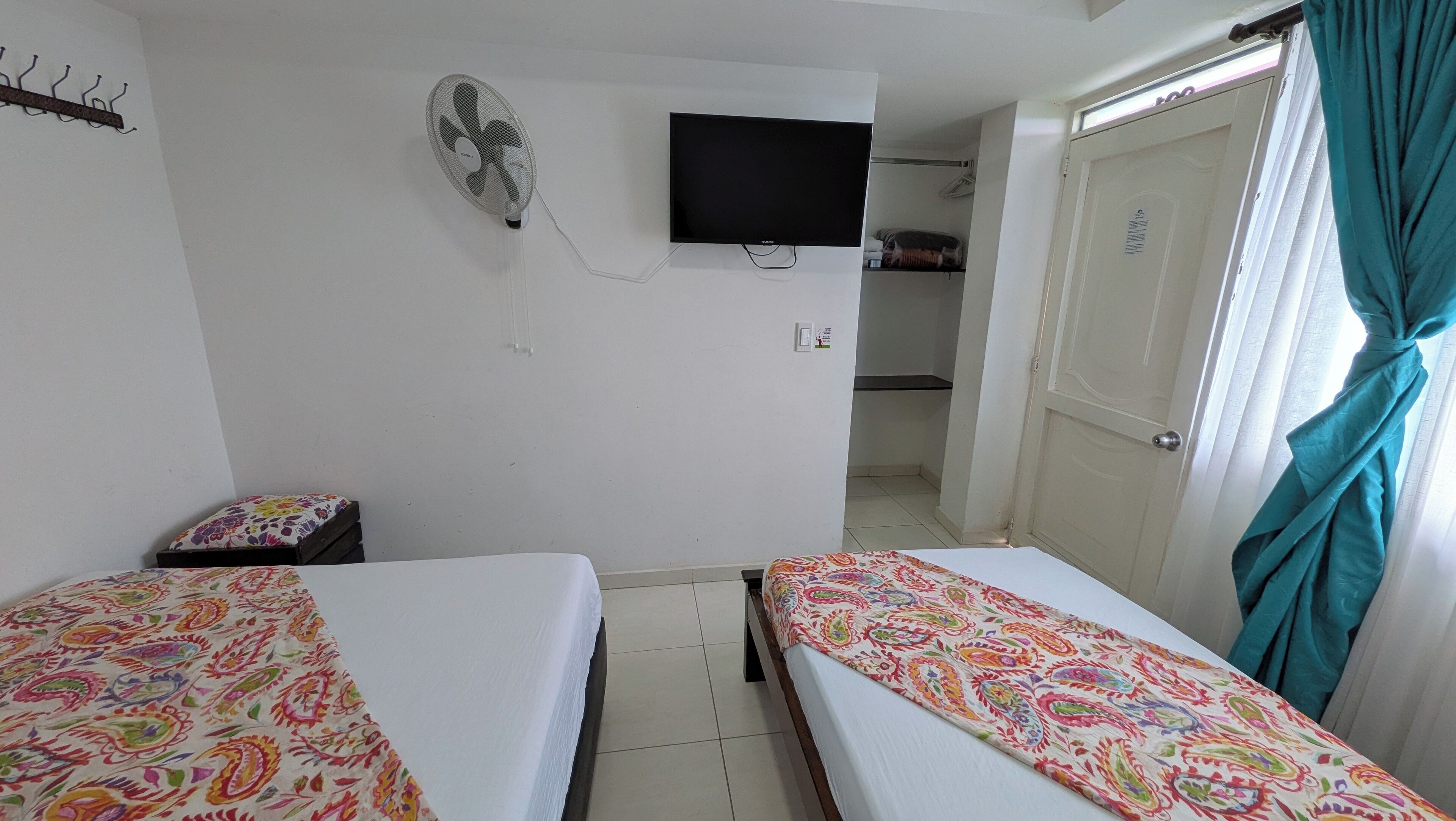 Een kamer met twee bedden, een aan de muur bevestigde televisie, een ventilator en een deur voorzien van gordijnen.