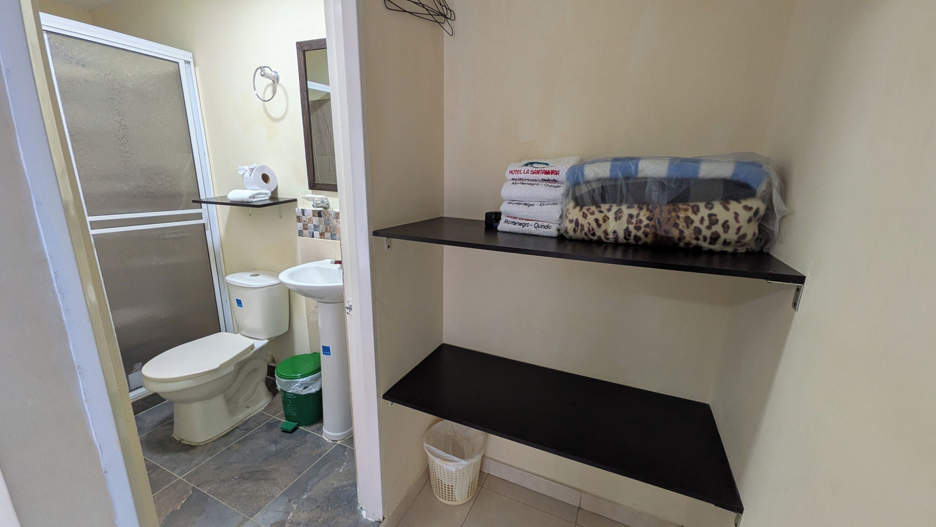 A bathroom with a toilet, sink, and a shower area. There are shelves with toiletries and a trash bin.