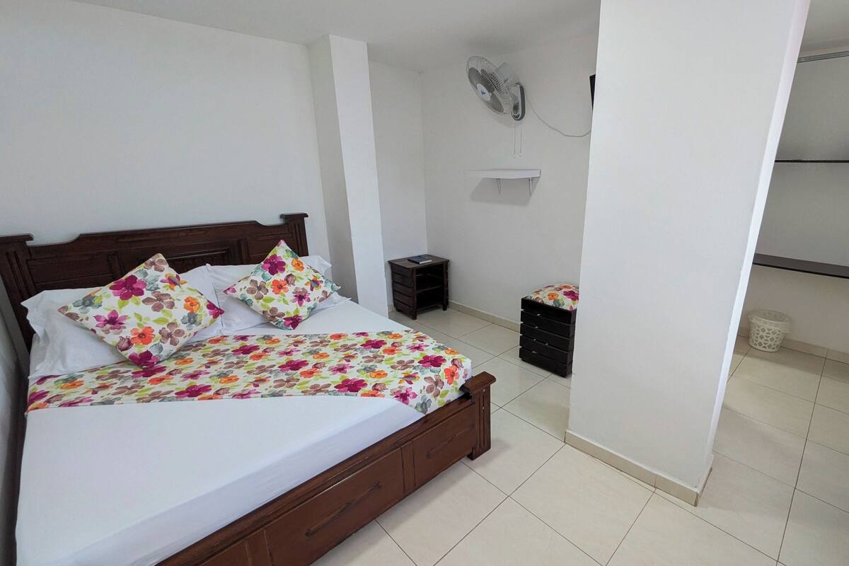 Une chambre à coucher bien rangée, avec un lit en bois, une literie à motifs floraux, une table de chevet, un petit tiroir, un ventilateur fixé au mur et une armoire dans laquelle est suspendu un sac.