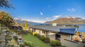 A modern house with a metal roof, a well-maintained lawn, and a view of a lake and mountains. - Panorama Terrace: Lake Views, Deck & Walk to Town (Queenstown)