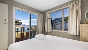 A hotel room with a large bed, a balcony with a view of mountains, and a wooden dresser. - Panorama Terrace: Lake Views, Deck & Walk to Town (Queenstown)