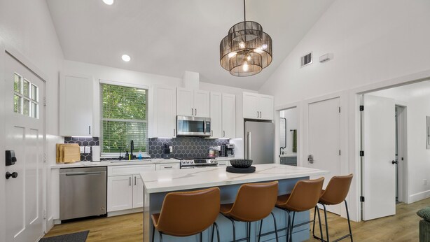 A modern kitchen with white cabinets, a large island with bar stools, a hanging light fixture, and a window with greenery outside.