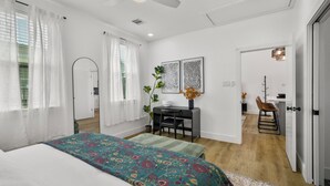 A bedroom with a bed, a desk, and a chair. There is a ceiling fan, a window with curtains, and a door leading to another room.