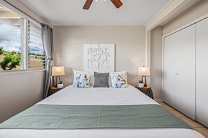 A bedroom with a bed, two bedside lamps, a window with curtains, a ceiling fan, and a wall-mounted artwork. - 2bd tropical, walk to beach stay (Honokowai)
