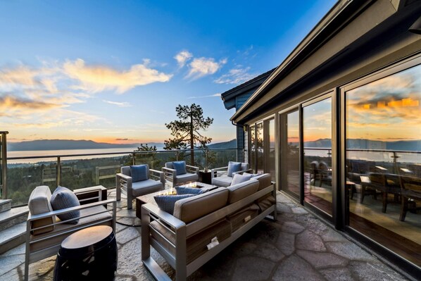 Un espace extérieur moderne aménagé avec des sièges, offrant une vue sur un lac et des montagnes au coucher du soleil.