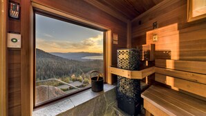 A wooden cabin interior with a large window offering a scenic view of a forest and mountains. A stone fireplace with a wooden bench is visible. - Tahoe Blue Estate Luxury Retreat LUXE5 (Stateline)