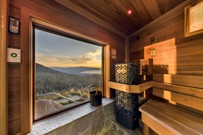 A wooden cabin interior with a large window offering a scenic view of a forest and mountains. A stone fireplace with a wooden bench is visible. - Tahoe Blue Estate Luxury Retreat LUXE5 (Stateline)