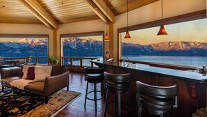 A cozy living area with a wooden ceiling, a sofa, a coffee table, and a bar with stools. Large windows offer a view of snow-capped mountains and a lake. - Tahoe Blue Estate Luxury Retreat LUXE5 (Stateline)