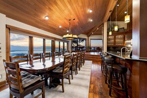 Un vaste espace dédié à la salle à manger, avec une grande table et des chaises en bois, un bar avec des tabourets, et de grandes fenêtres offrant une vue sur les montagnes.