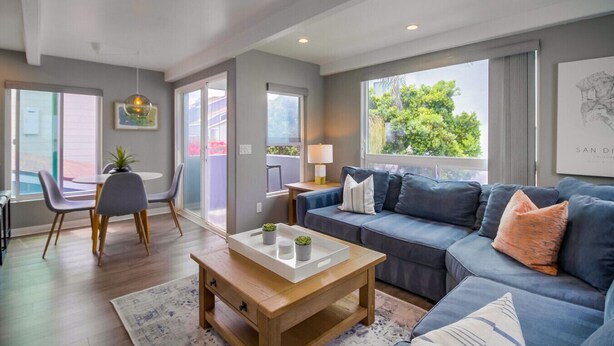 A modern living room with a sofa, coffee table, and chairs. There is a sliding glass door leading to an outdoor area. - A-Wave From It All, Steps to Sand with AC! (San Diego)