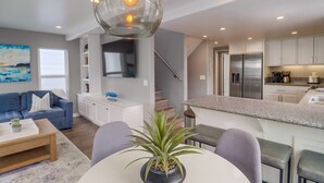 A modern living room with a blue sofa, a coffee table, and a potted plant. There is a kitchen area with granite countertops and stainless steel appliances. - A-Wave From It All, Steps to Sand with AC! (San Diego)