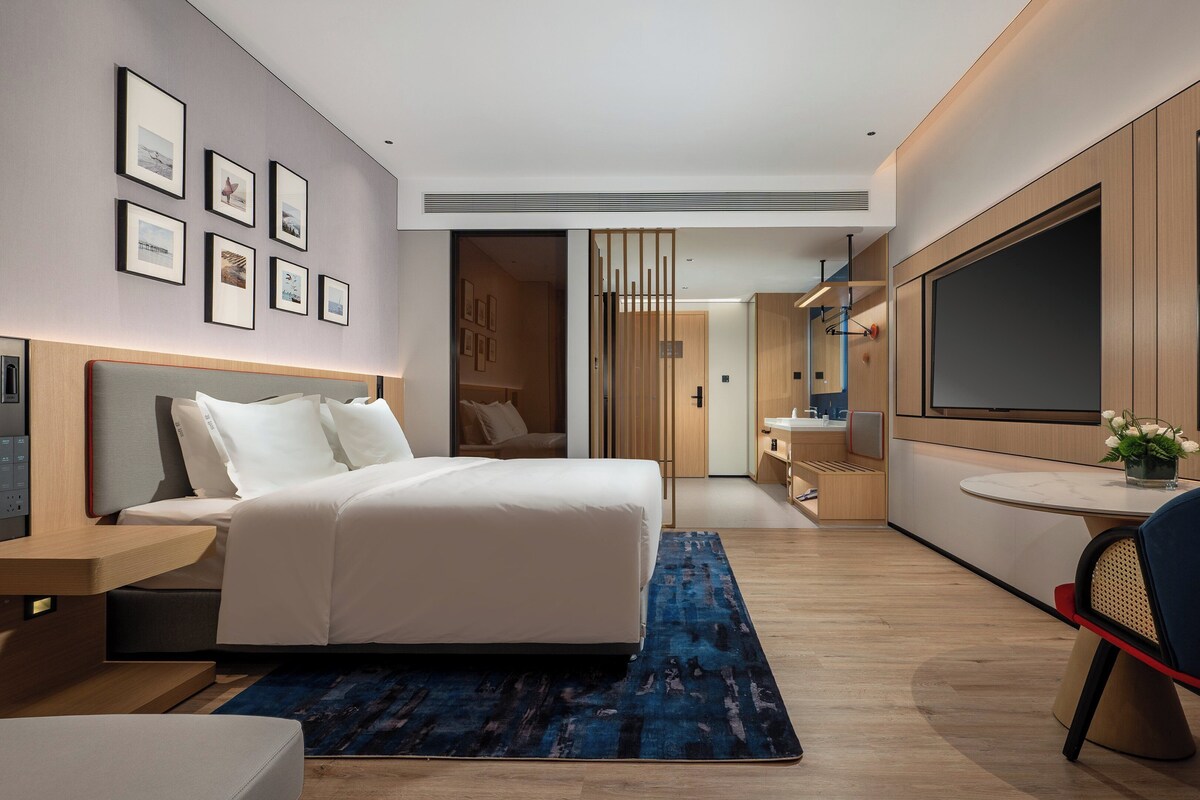A modern hotel room with a large bed, bedside tables, a desk, a chair, a TV, and framed pictures on the wall.